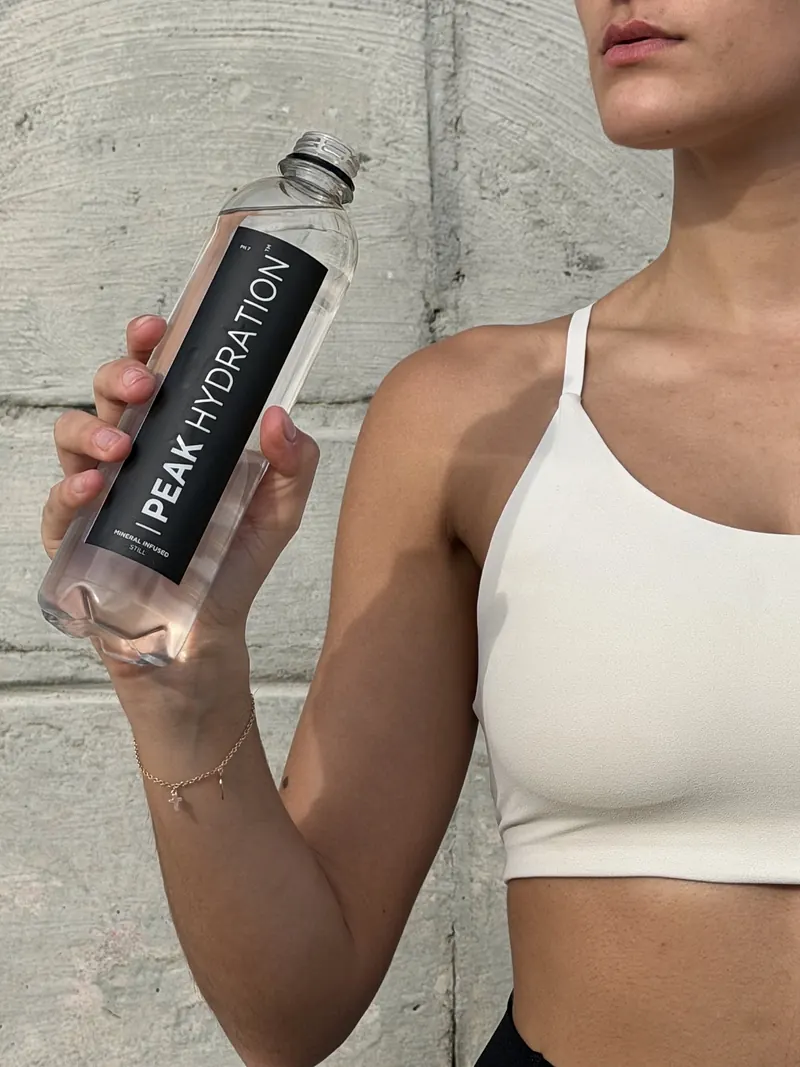A woman holding a bottle of Peak Hydration still water against a concrete wall